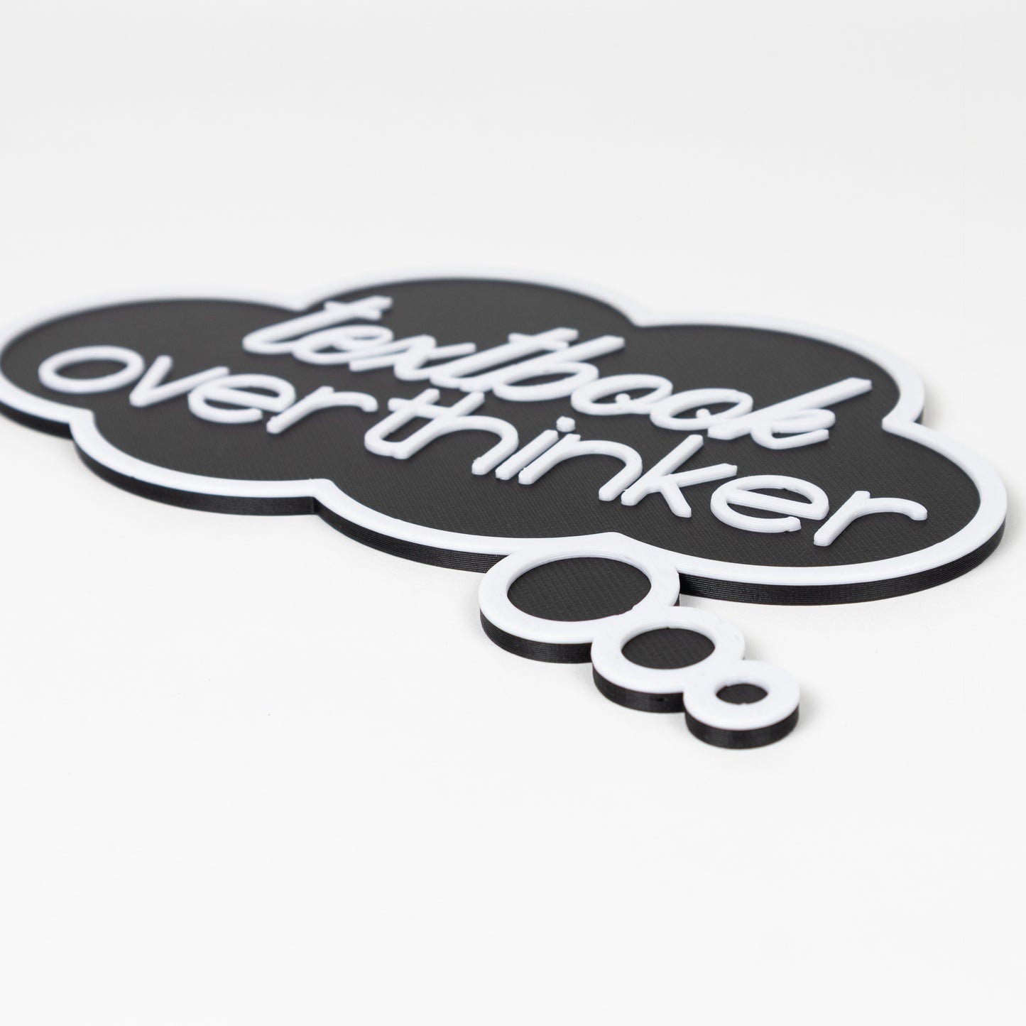 Close up of black thought bubble sign with raised white text that says, 'textbook overthinker'. Photo booth prop shot on a white background.