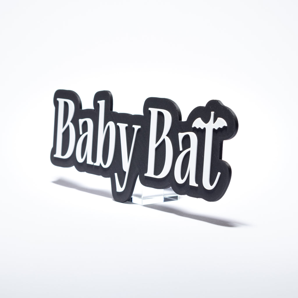 A black 3d printed sign featuring the words 'Baby Bat' in white script with a small bat silhouette integrated into the lettering, displayed on a clear acrylic stand against a white background.