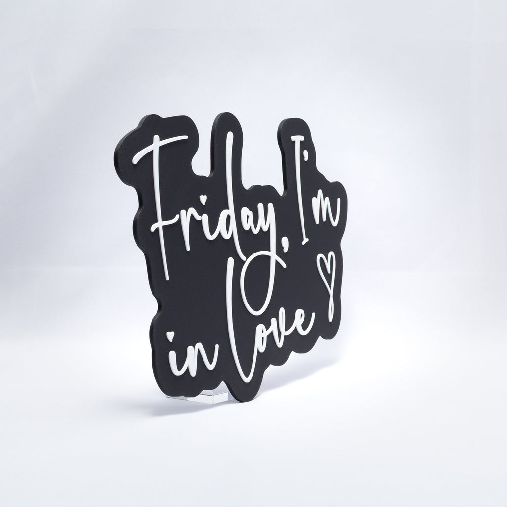 A 3D printed sign with white text on a black base that says, "Friday, I'm In Love" with a heart at the end. Shot at an angle on a white background.