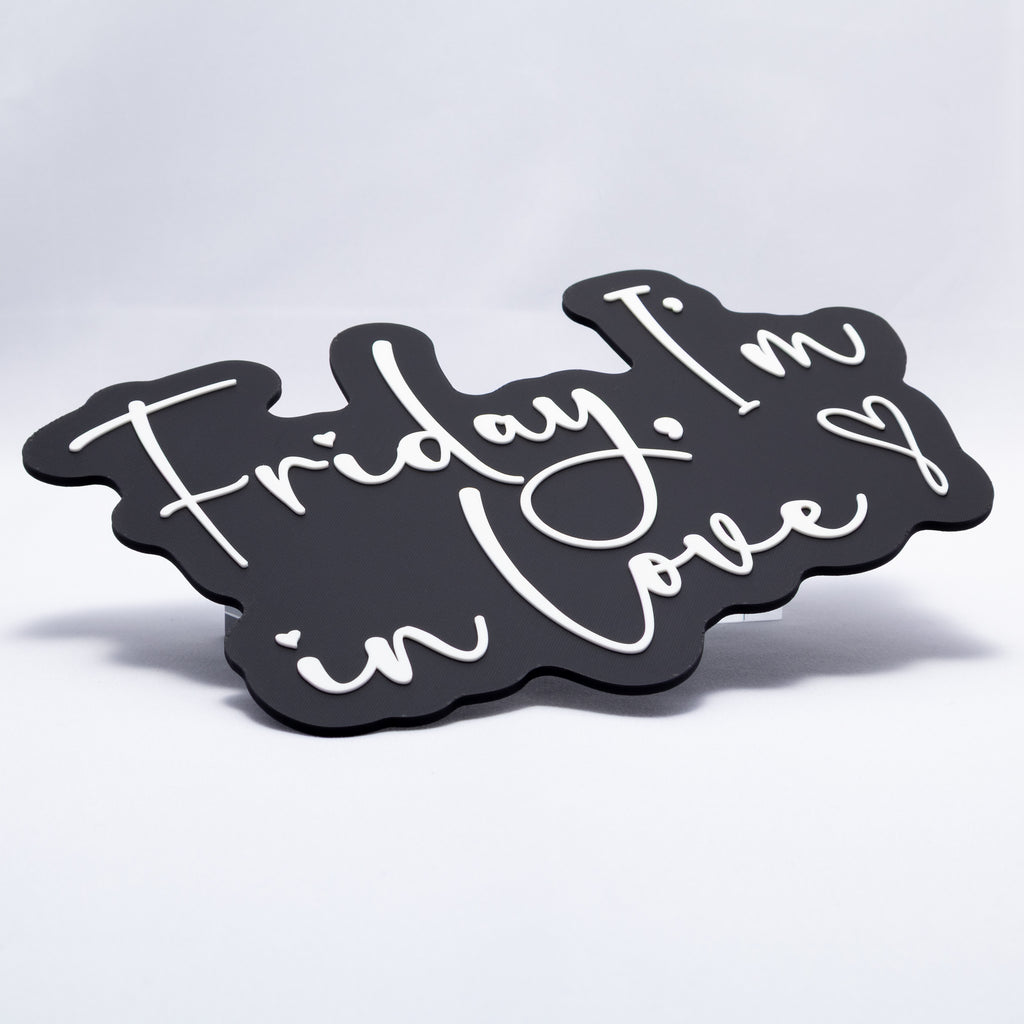 A 3D printed sign with white text on a black base that says, "Friday, I'm In Love" with a heart at the end. Shot almost flat to show raised texture of white lettering.