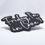 A 3D printed sign with white text on a black base that says, "Friday, I'm In Love" with a heart at the end. Shot almost flat to show raised texture of white lettering.