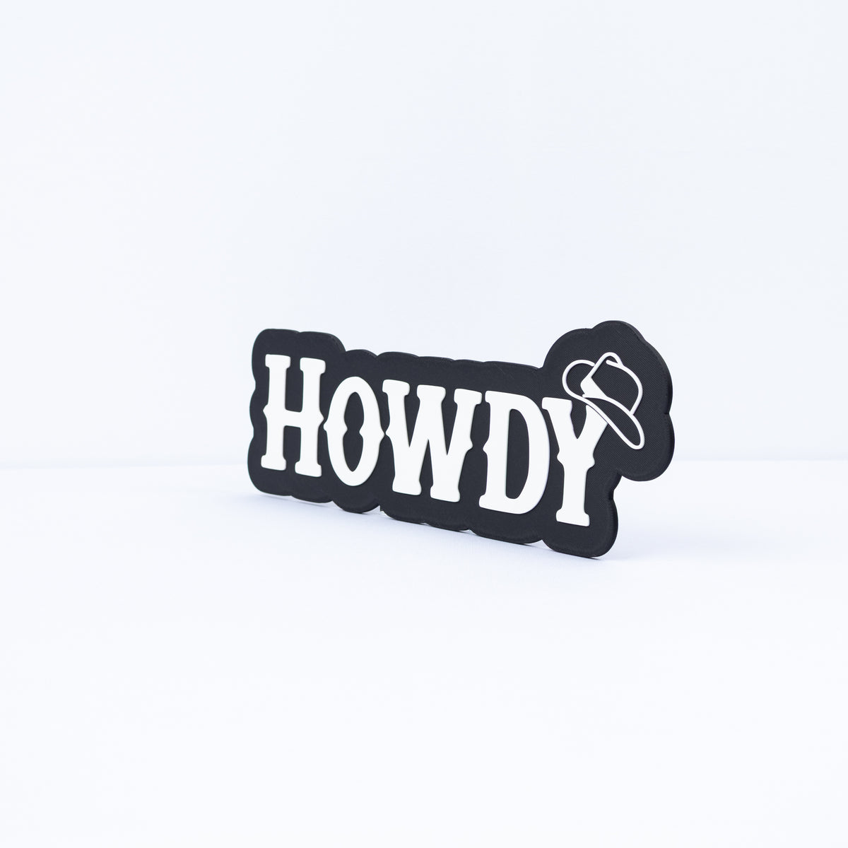 A 3D printed black sign with white Western style lettering that says, "Howdy". A cowboy hat hangs on the end of the letter "y". Shot at an angle.