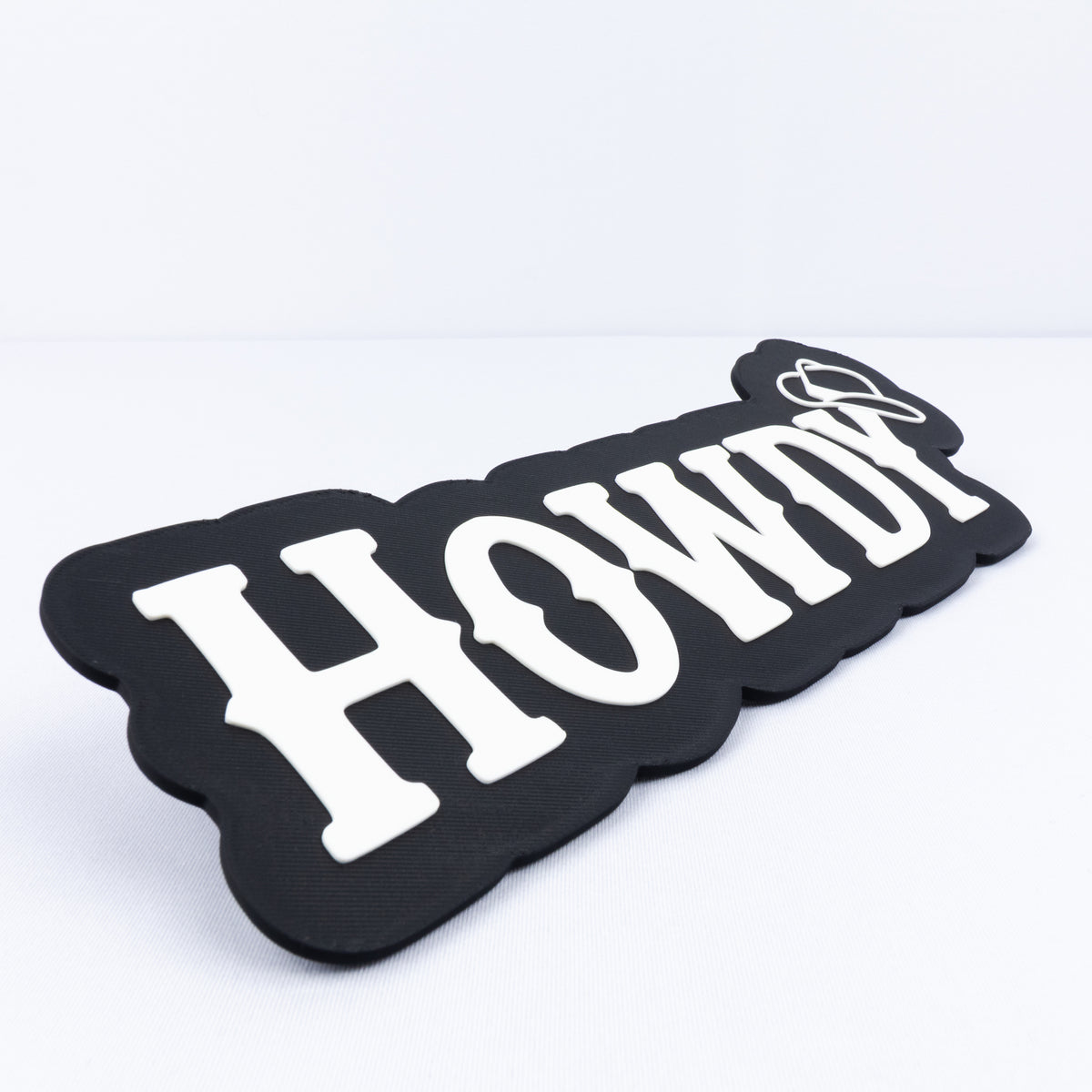 A 3D printed black sign with white Western style lettering that says, "Howdy". A cowboy hat hangs on the end of the letter "y". Close up shot to show raised white lettering.