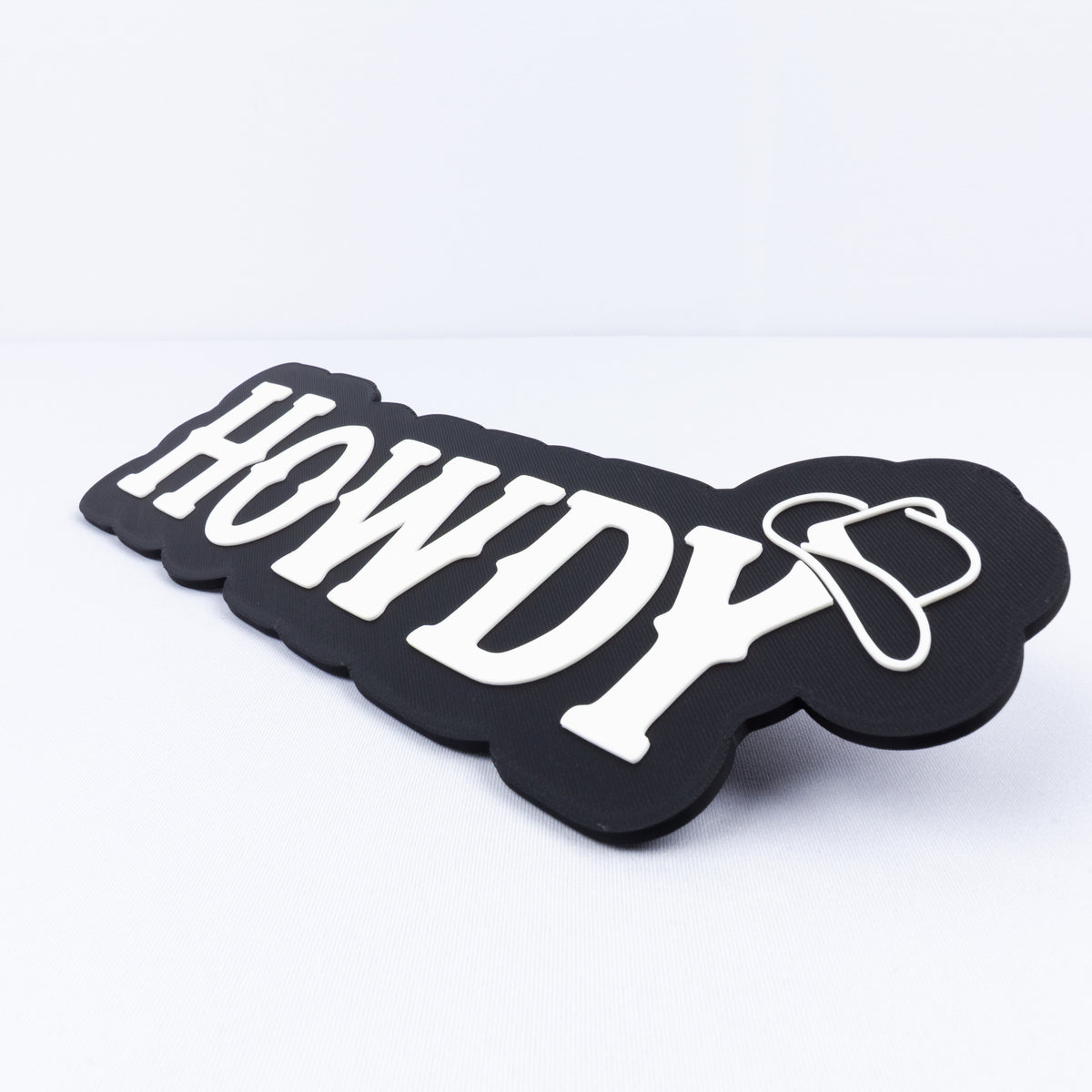 A 3D printed black sign with white Western style lettering that says, "Howdy". A cowboy hat hangs on the end of the letter "y". Close up shot to show raised white lettering and cowboy hat.