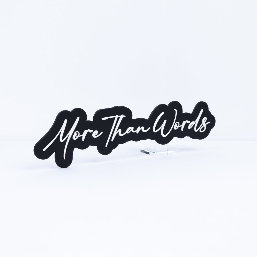 A black 3d printed sign with the song title 'More Than Words' in white cursive script, displayed on a small clear acrylic stand against a clean white background.