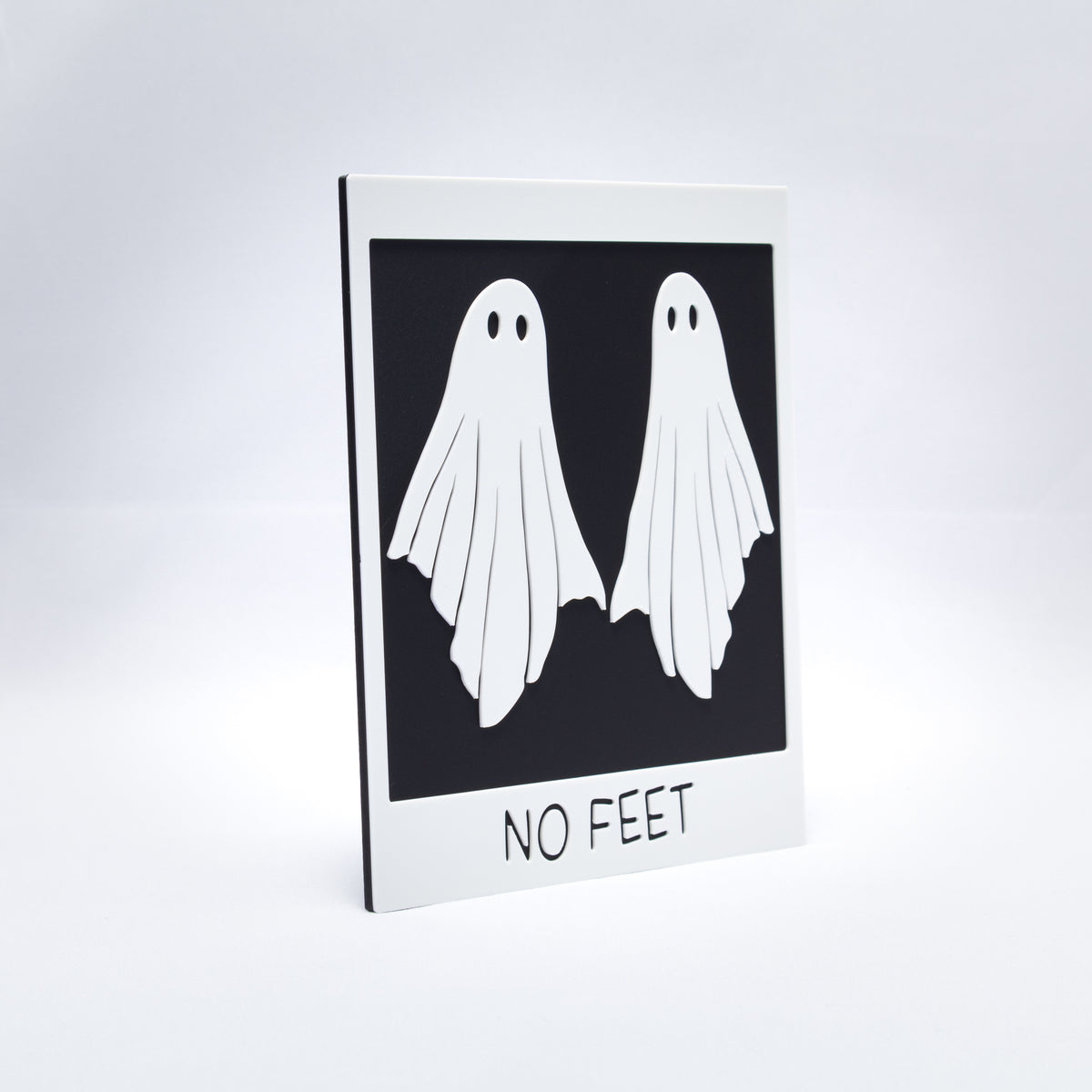A 3D printed black and white sign that looks a polaroid photo of two ghosts with the words, "No Feet" at the bottom. Shot at an angle to show texture.