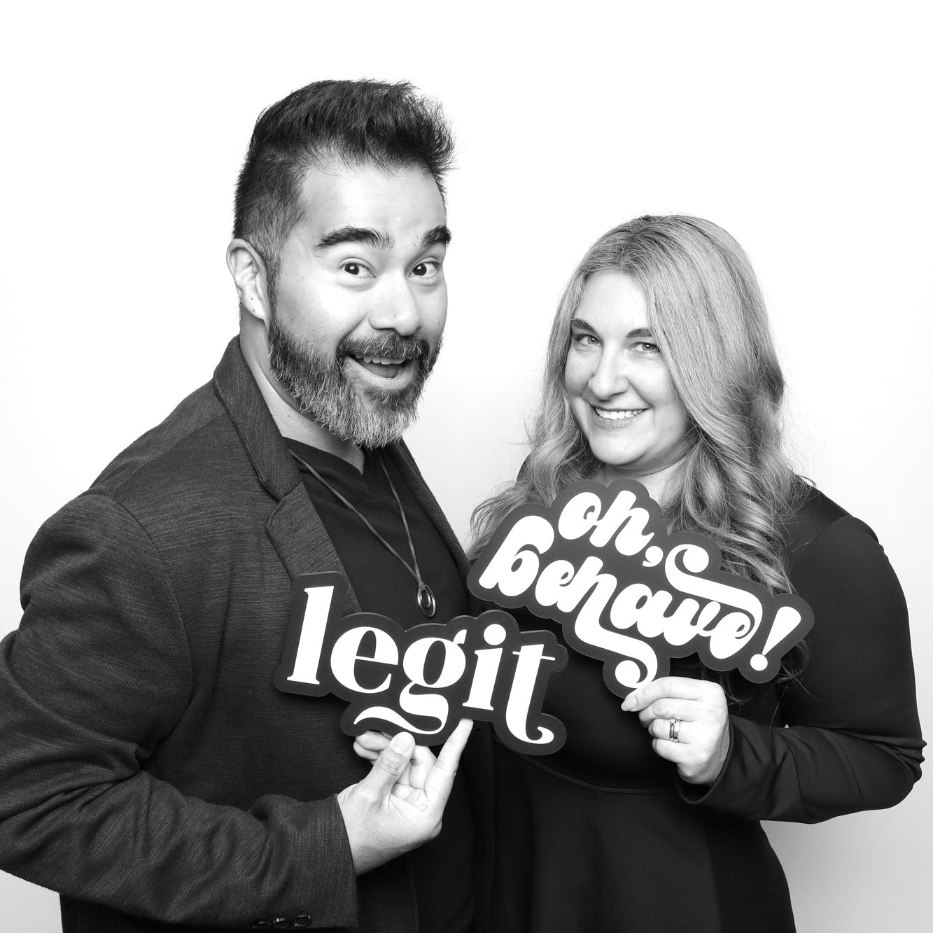 Two people holding signs that say, "Oh Behave!" and "legit". Posing against a white background in a photo booth.