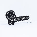 A black decorative sign featuring the word 'Starman' in white script with small white stars, displayed on a clear acrylic stand against a clean white background.