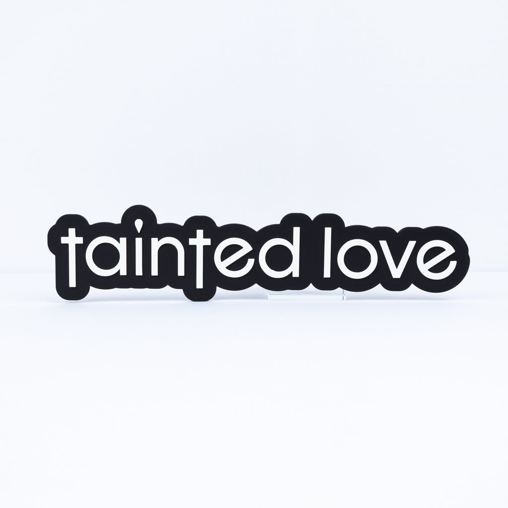 A black sign with the words 'tainted love' in white lowercase letters, for music themed parties and photo booth events.