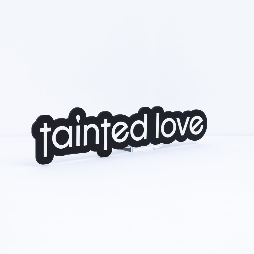 A black sign with the words 'tainted love' in white lowercase letters, for music themed parties and photo booth events.