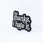A funny black decorative sign with white script text reading 'thanks, i hate it.' displayed on a small clear stand against a clean white background.