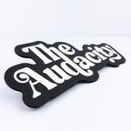 A black sign prop with the words 'The Audacity' in raised white retro-style lettering, for use at parties and photo booth events.