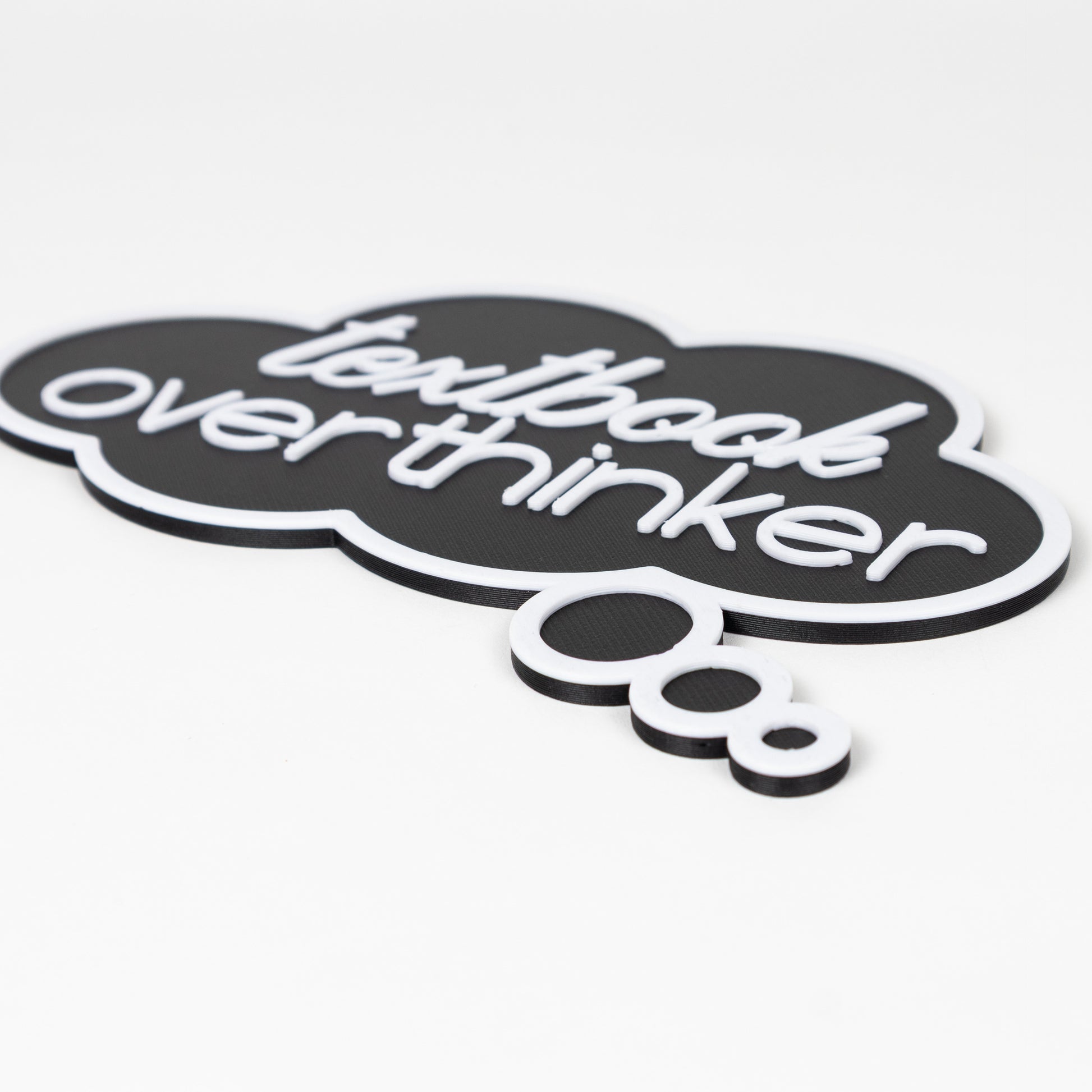 Close up of black thought bubble sign with raised white text that says, 'textbook overthinker'. Photo booth prop shot on a white background.